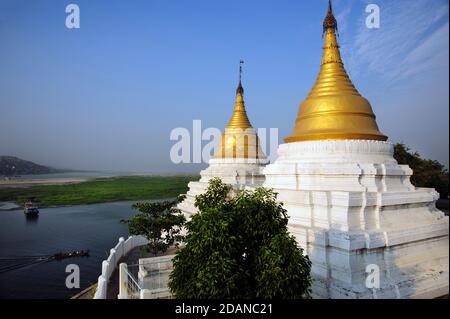 Ein burmesisches Boot passiert zwei vergoldete burmesische Tempelstupas Am Ufer des Ayeyarwady Flusses mit Blick auf Sagaing in der Nähe von Mandalay Myanmar Stockfoto