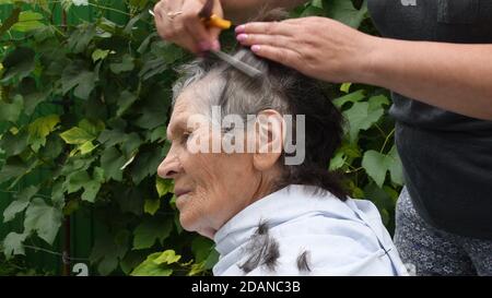 Ältere Frau Profil Porträt beim Schneiden ihrer Haare zu Hause im Hinterhof im Freien. Verschwommene weibliche Hände kämmen graue Haare von 80 Jahre alte Frau. Wrinkl Stockfoto