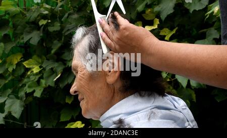 Nahaufnahme des Haarschneidens von älteren Frauen zu Hause im Hinterhof im Freien. Zu Hause bleiben während der Quarantäne. Frau Hände halten Kamm und Schere zu Stockfoto