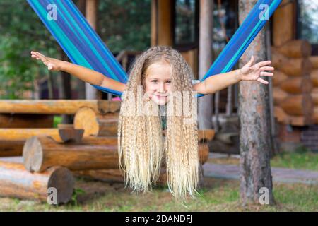 Kleines glückliches Mädchen mit langen blonden lockigen Haaren schwingt die Arme auf einer blau-grünen Hängematte ausgestreckt. Bewegungsfreiheit, Lebensstil. Schulferien, Urlaub Stockfoto