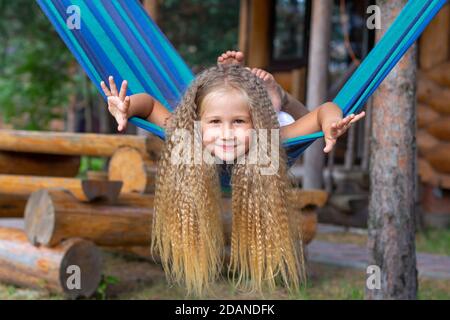 Kleines glückliches Mädchen mit langen blonden lockigen Haaren schwingt die Arme auf einer blau-grünen Hängematte ausgestreckt. Bewegungsfreiheit, Lebensstil. Schulferien, Urlaub Stockfoto