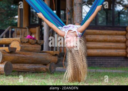 Kleines glückliches Mädchen mit langen blonden lockigen Haaren schwänzt kopfüber auf einer blau-grünen Hängematte. Bewegungsfreiheit, Lebensstil. Schulferien, Urlaub, Tag Stockfoto