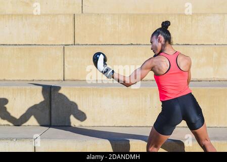 Junge afroamerikanische Boxerin trägt rosa und schwarz Sport Kleidung und schwarze Boxhandschuhe, die mit einem linken Haken stanzen In einem Park und projizieren s Stockfoto