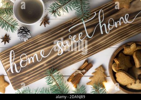 Home Sweet Home Schriftzug verbranntes Holzschild, Lebkuchenkeks, Christbaumzweige, Tasse Kaffee, Zimtstangen auf weißer Oberfläche. Flach liegend. Stockfoto