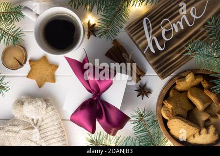 Startseite Schriftzug verbranntes Holzschild, Lebkuchenkeks, Christbaumzweige, Tasse Kaffee, Geschenk, Zimtstangen auf weißer Oberfläche. Flach liegend Stockfoto