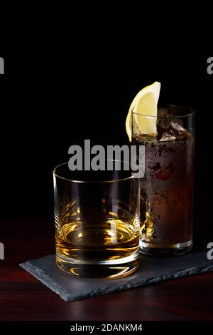 Whiskey ordentlich und Cola mit Eis und Zitronenkeil serviert auf Schieferplatte. Dunkelroter Holztisch, hohe Auflösung Stockfoto