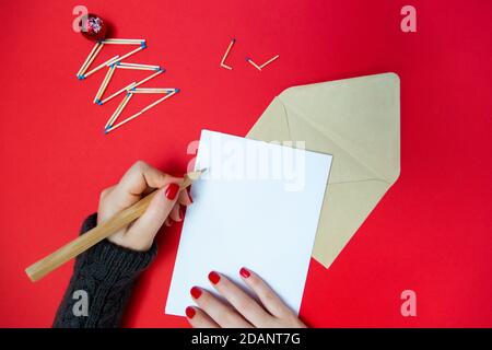 Die Hand des Mädchens in einem Pullover schreibt einen Brief auf den Hintergrund eines Weihnachtsbaums aus Streichhölzern. Roter Hintergrund. Stockfoto