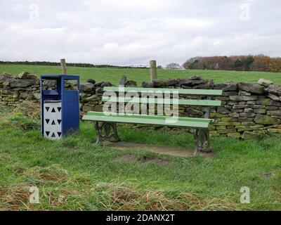 Grün und schwarz Parkbank grau und blau Abfalleimer Stand auf Gras vor der Trockensteinmauer mit Bäume und Felder im Hintergrund Stockfoto