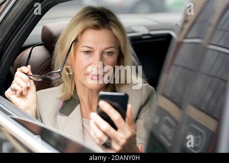 Blonde ernsthafte reife Frau lesen Nachricht im Smartphone auf dem Rücksitz Des Autos Stockfoto