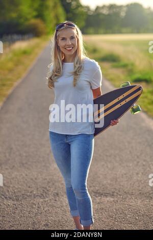 Porträt einer fit schöne Frau mittleren Alters mit einem aktiven Lebensstil lächeln und Blick auf die Kamera, während ein Longboard auf einer sonnigen Straße in der Stockfoto