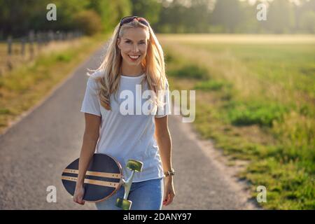 Porträt einer fit schöne Frau mittleren Alters mit einem aktiven Lebensstil lächeln und Blick auf die Kamera, während ein Longboard auf einer sonnigen Straße in der Stockfoto