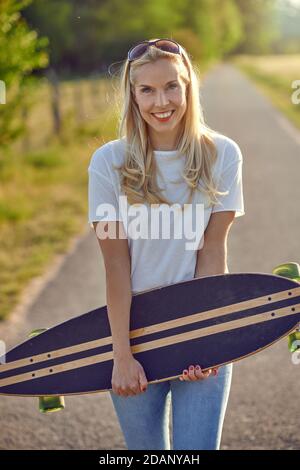 Porträt einer fit schöne Frau mittleren Alters mit einem aktiven Lebensstil lächeln und Blick auf die Kamera, während ein Longboard auf einer sonnigen Straße in der Stockfoto