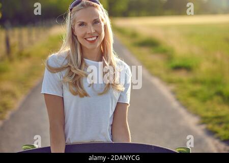 Porträt einer fit schöne Frau mittleren Alters mit einem aktiven Lebensstil lächeln und Blick auf die Kamera, während ein Longboard auf einer sonnigen Straße in der Stockfoto