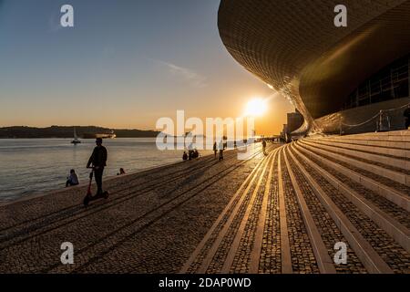 Die Silhouetten von Menschen und Schiffen bewegen sich auf dem Tejo und auf dem PAVE Walk neben Maat, Museum für Kunst, Architektur und Technologie in Lissabon, Portugal Stockfoto