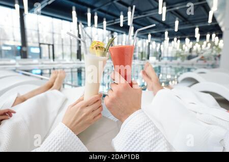 Hände von jungen erholsamen Paar in weißen Bademänteln klirren mit Cocktails Stockfoto