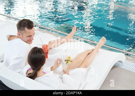Glückliches junges erholsames Paar in weißen Bademänteln, die sich unterhalten und haben Cocktails Stockfoto