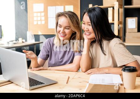 Glückliche junge asiatische Frau am Tisch neben Kollegen sitzen Vor dem Laptop Stockfoto