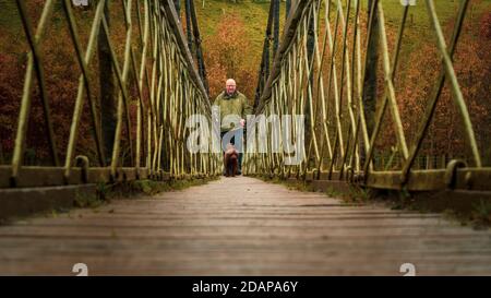 Walker und Hund überqueren die Hebden Hängebrücke auf dem Dales Way über den Fluss Wharfe im Herbst, Yorkshire Dales National Park, UK Landschaft Stockfoto