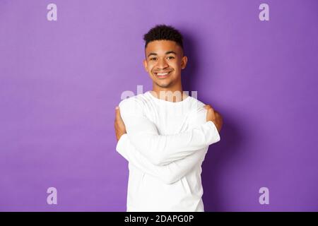 Fröhlicher afroamerikanischer Mann in weißem Sweatshirt, umarmt sich und lächelt, glücklich über purpurem Hintergrund stehend Stockfoto