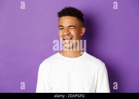 Nahaufnahme eines hübschen jungen afroamerikanischen Mannes, der blinzelnd, lächelnd und fröhlich aussagt und auf purpurem Hintergrund steht Stockfoto