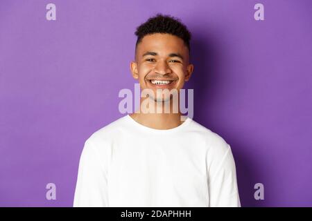 Nahaufnahme eines jungen afroamerikanischen Mannes, lächelnd zufrieden, zufrieden stehend auf violettem Hintergrund, mit weißem Sweatshirt Stockfoto