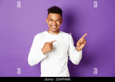 Bild eines gutaussehenden und sorglosen afroamerikanischen Mannes in weißem Sweatshirt, mit den Fingern nach rechts zeigend und über etwas Lustiges lachend, über Purpur stehend Stockfoto
