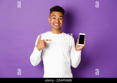 Portrait von schönen afroamerikanischen Mann, lächelnd und Zeigefinger auf Smartphone-Bildschirm, zeigt Anwendung, stehen über lila Hintergrund Stockfoto