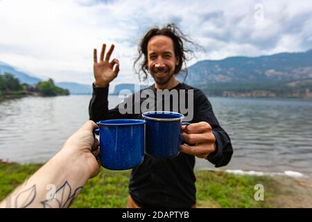 Selektiver Fokus einer tätowierten Person, die eine Tasse Kaffee oder Tee einklinkt, mit einem sehr glücklichen jungen Mann, während er vor einem See und Bergen steht Stockfoto