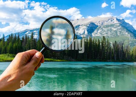 Eine Nahaufnahme einer Hand, die eine Lupe über einer ruhigen Berglandschaft hält, auf der Suche nach Reise- und Campingplätzen in Alberta, Kanada Stockfoto