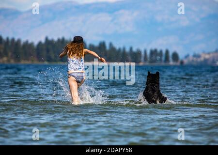 Langhaarige Frau und ihr Hund schossen von hinten, rannten vor der Kamera und spritzte knietief in das Wasser eines kanadischen Sees, aufsteigende Spritzer. Stockfoto