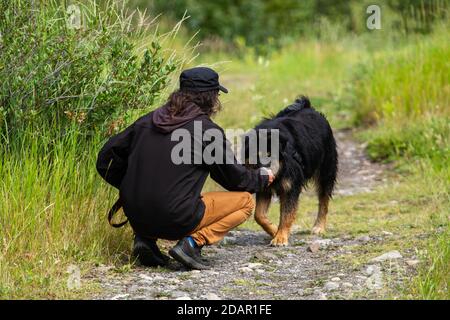 Mann mit langen Haaren und schwarzem Hut und Jacke von hinten geschossen, kniend, um seinen großen schwarzen berner aussehenden Hund bei einem Spaziergang auf einem Bergpfad streicheln. Stockfoto