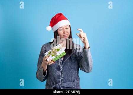 Enttäuscht asiatische junge Frau in Weihnachtsmann Hut hält und Zeigt ihren Fingern eine kleine Geschenkbox auf einem Blauer Hintergrund Stockfoto