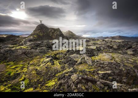 Mann auf einem Hügel in einem schwarzen Lavafeld mit grünem Moos, das Hände in den dramatischen Himmel streckt, Leihrnjukur in der Krafla, Skutustaoir, Norourland Stockfoto