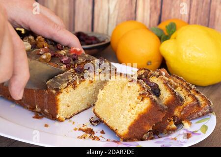 Die Hände des Mannes schneiden einige Scheiben einer hausgemachten Zitrone und Orangenpudding mit Früchten auf einer weißen Platte im Vintage-Stil Blumenzeichnungen auf einem hölzernen t Stockfoto