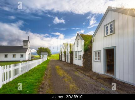 Weiße Häuser mit Grasdach und Steinmauer entlang einer Schotterstraße, Kirche mit weißem Zaun auf Torfhof, Grashaus Siedlung Grenjadarstadur Stockfoto