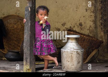 Ein kleines Mädchen steht vor dem Haus neben einem Aluminium-Wasserbehälter und kaut Zuckerrohr, Manaslu-Region, Himalaya, Nepal Stockfoto