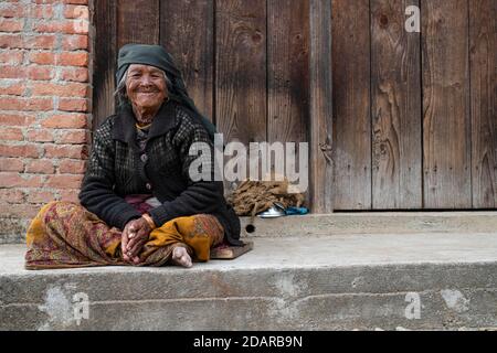 Freundliche lächelnde ältere Frau mit Kopftuch und Nasenring sitzt vor einer Holztür, Himalaya, Jiri, Khumbu Region, Nepal Stockfoto