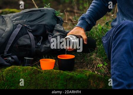 Tee trinken während der Wanderung. Mann Hand Gießen heißen Tee aus schwarzen Thermosflasche in 2 Tassen im Wald. Erfrischung beim Wandern. Campingausrüstung Stockfoto