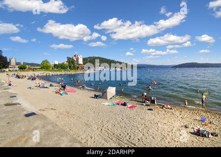 Touristen und Einheimische Idahoans entspannen sich am sandigen Stadtstrand entlang des Lake Coeur d'Alene mit dem Resort und dem Tubbs Hill im Hintergrund. Stockfoto