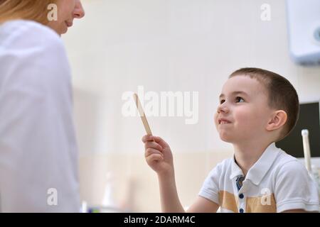 Der Arzt und der Patient sind ein Kind. Der Junge hält einen Spatel in der Hand und sieht wehmütig lächelnd aus. Stockfoto