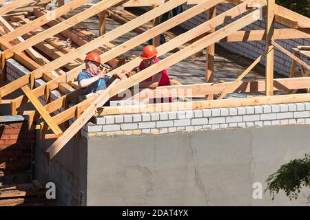 GRODNO, WEISSRUSSLAND - JUNI 2020: Zwei Arbeiter Dachdecker in Helmen umgeben von Sparren und Balken auf der Baustelle bauen Haus. Tischlerset Stockfoto