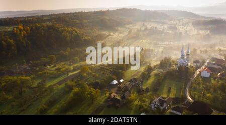 Luftaufnahme des Dorfes im Nebel mit goldenen Sonnenstrahlen bei Sonnenaufgang im Herbst. Schöne ländliche Landschaft mit nebligen bunten Bäumen, Kirche, orange Himmel mit Stockfoto