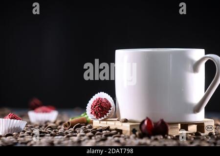 Weiße Tasse voller Kaffeebohnen, roten Blumen und roten Schokolade Süßigkeiten vor dem schwarzen Hintergrund. Espresso am Morgen. Kaffeetasse. Stockfoto
