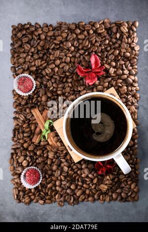 Flache Ansicht einer weißen Tasse voller Kaffeebohnen in Form eines Fotorahmens mit roten Schokoladenbonbons und roten Blumen. Espresso am Morgen. Kaffeetasse. Stockfoto