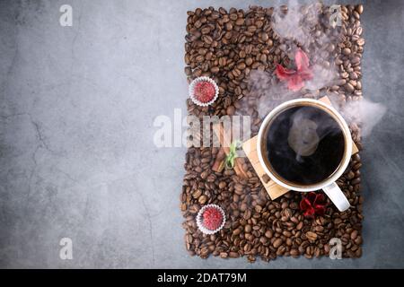 Flache Ansicht einer weißen Tasse voller Kaffeebohnen in Form eines Fotorahmens mit roten Schokoladenbonbons und roten Blumen. Espresso am Morgen. Kaffeetasse. Stockfoto