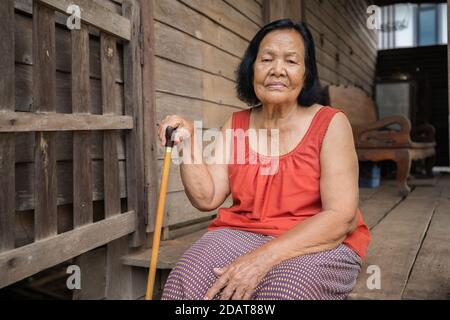 Thai ältere Frau in Rundhals ärmellose Kragen sitzt einsam in Altes Holzhaus Stockfoto