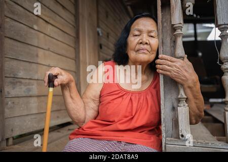 Thai ältere Frau in Rundhals ärmellose Kragen sitzt einsam in Altes Holzhaus Stockfoto