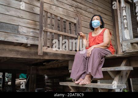 Thai ältere Frau in rund-Hals ärmellose Kragen trägt medizinische Maske Zum Schutz Corona Virus (covid-19) Pandemie in alten Holzhaus Stockfoto