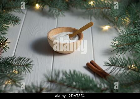 Zucker in einer Holzschüssel mit einem Löffel und einem Zimtstange neben grünen Zweigen eines Weihnachtsbaums Mit LED-Glühbirnen Girlande auf einem weißen Holz-Bac Stockfoto