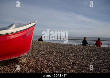 East Preston Kiesstrand an einem kühlen, aber sonnigen Herbsttag mit Einheimischen genießen den ruhigen Tag mit einem roten Boot im Vordergrund. Stockfoto